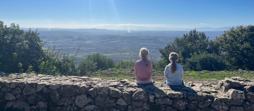 Zwei Kinder sitzen auf einer Steinmauer und blicken in eine Landschaft unter blauem Himmel.