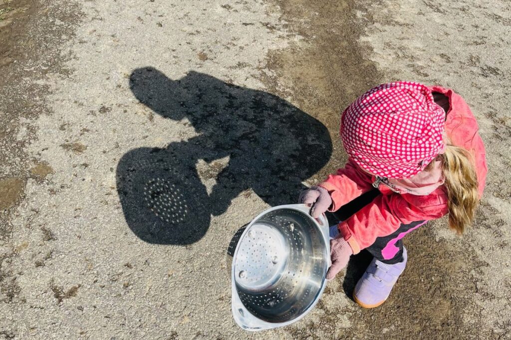 Kind beobachtet Lichtmuster der Sonne mit einem Küchensieb auf dem Boden