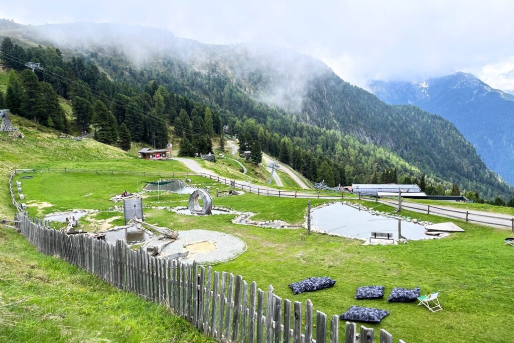 Blick auf den Zirbenpark am Hochzeiger im Pitztal mit Spiel- und Wasserbereichen vor der Tiroler Bergkulisse.