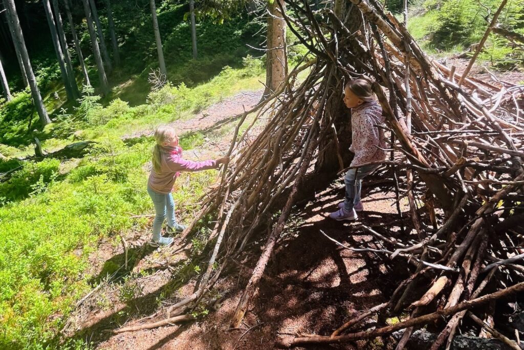 Zwei Kinder erkunden eine aus Ästen gebaute "Waldhütte" am Naturparkhaus Kaunergrat im Pitztal.